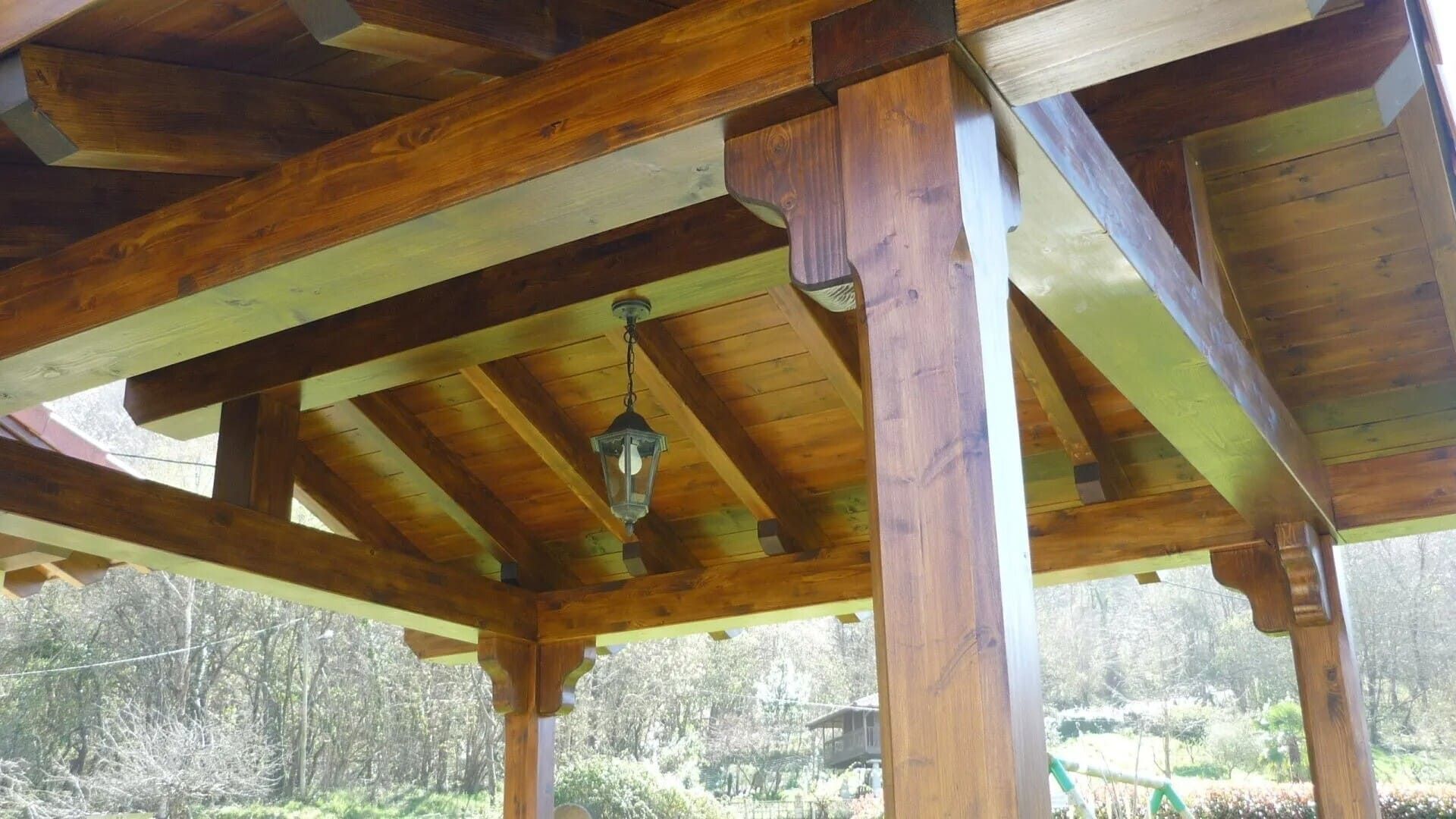 Gazebo de madera con techo a dos aguas y un farol colgante, visto desde abajo en un entorno boscoso al aire libre.