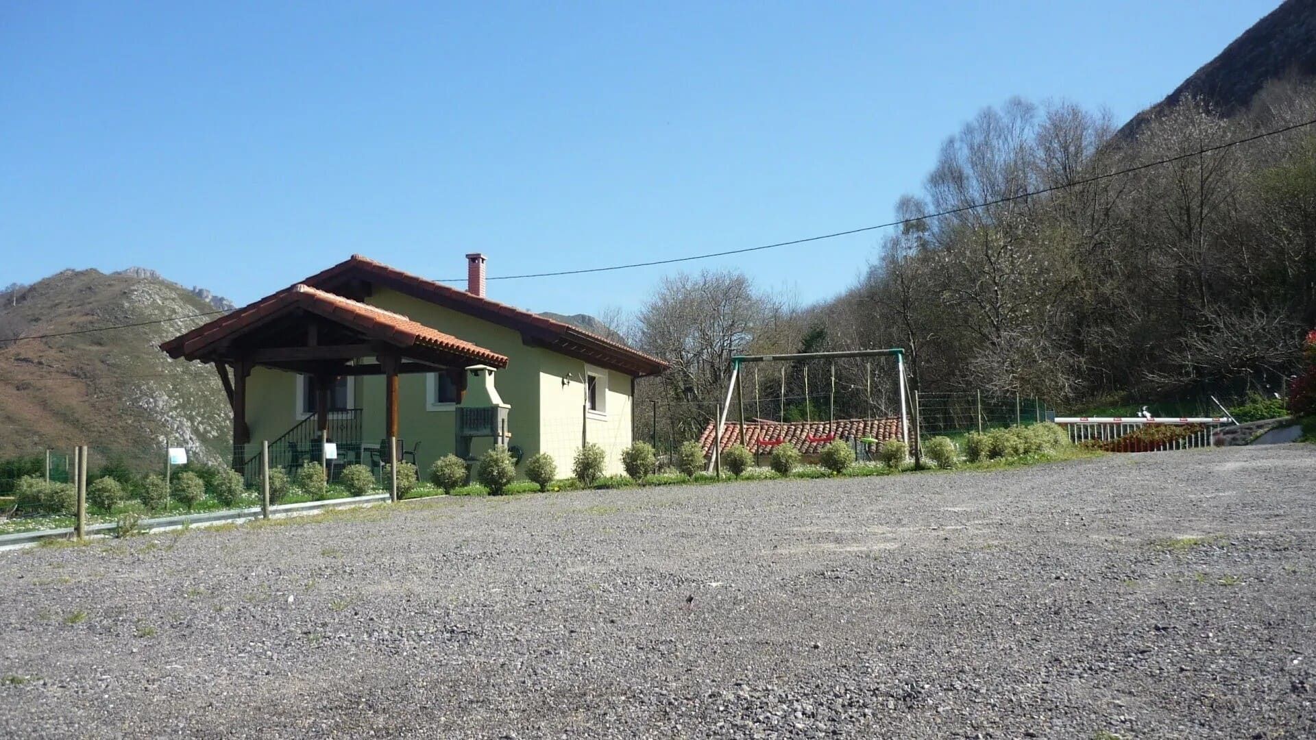 Casa pequeña con techo rojo junto a un terreno de grava, con árboles y colinas bajo un cielo azul despejado.
