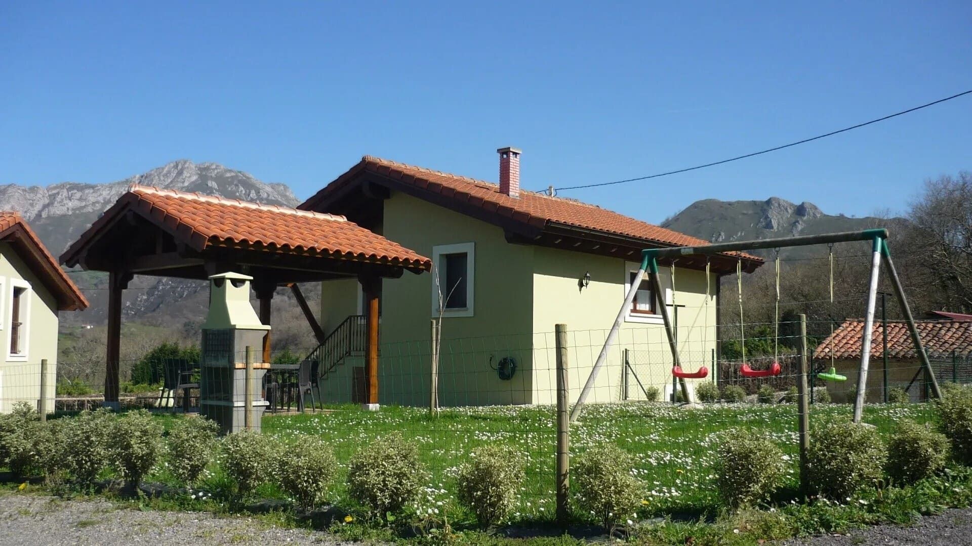 Casa rural con tejado de tejas rojas, paredes verdes y un jardín delantero, con las montañas y el cielo azul de fondo