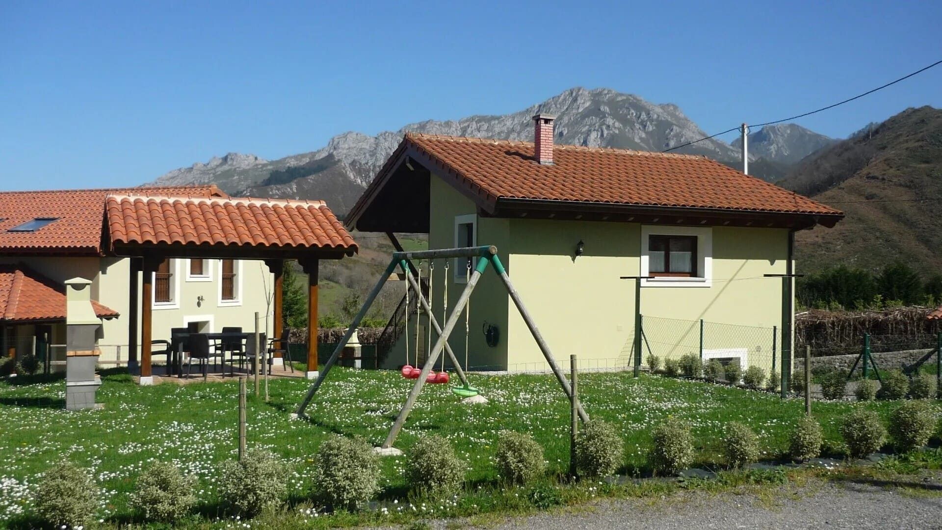 Dos casas pequeñas con tejados de tejas rojas junto a un jardín y montañas bajo un cielo despejado