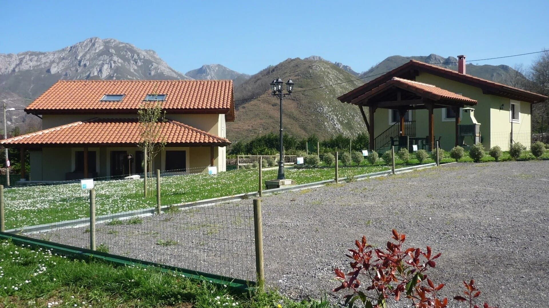 Dos casas con tejados rojos junto a un terreno de grava, con montañas y árboles al fondo.