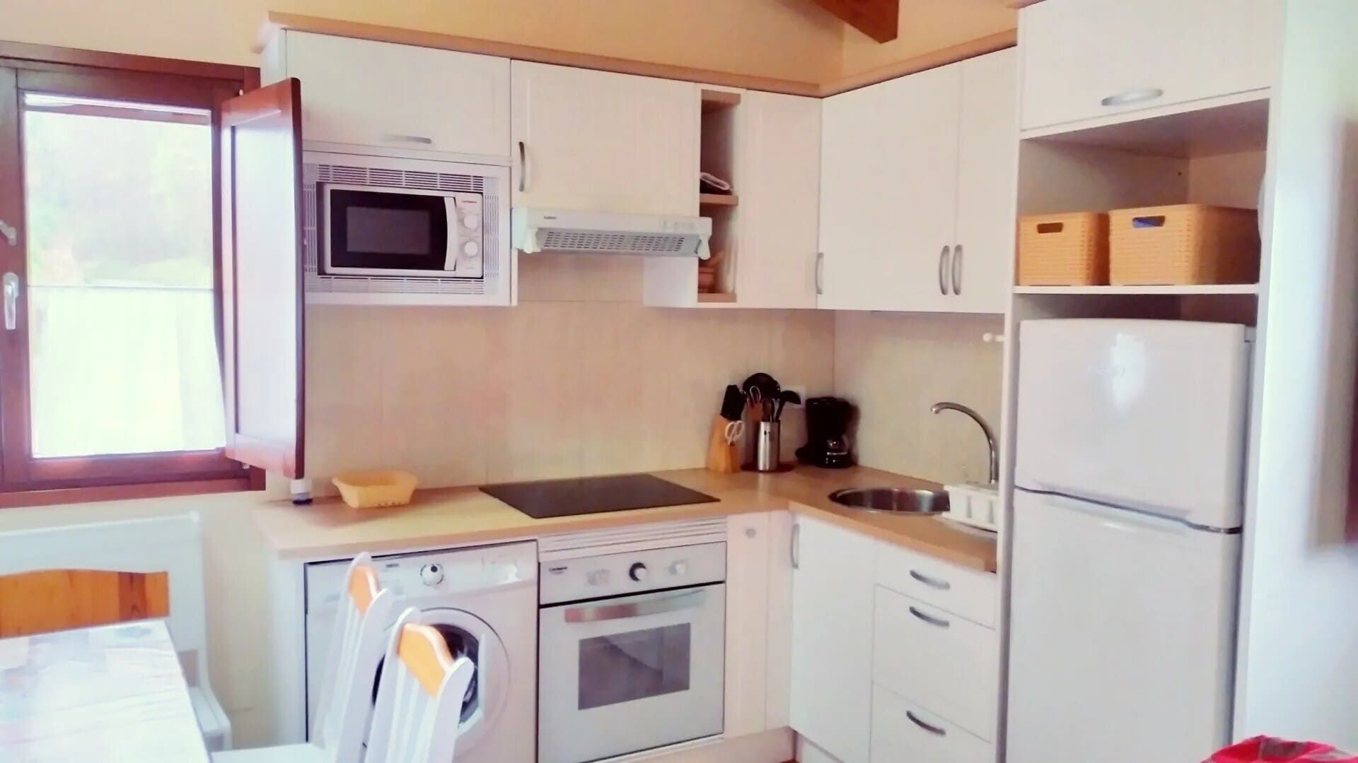Cocina compacta con armarios blancos, cocina, microondas, nevera y una ventana junto a una pequeña mesa de comedor.