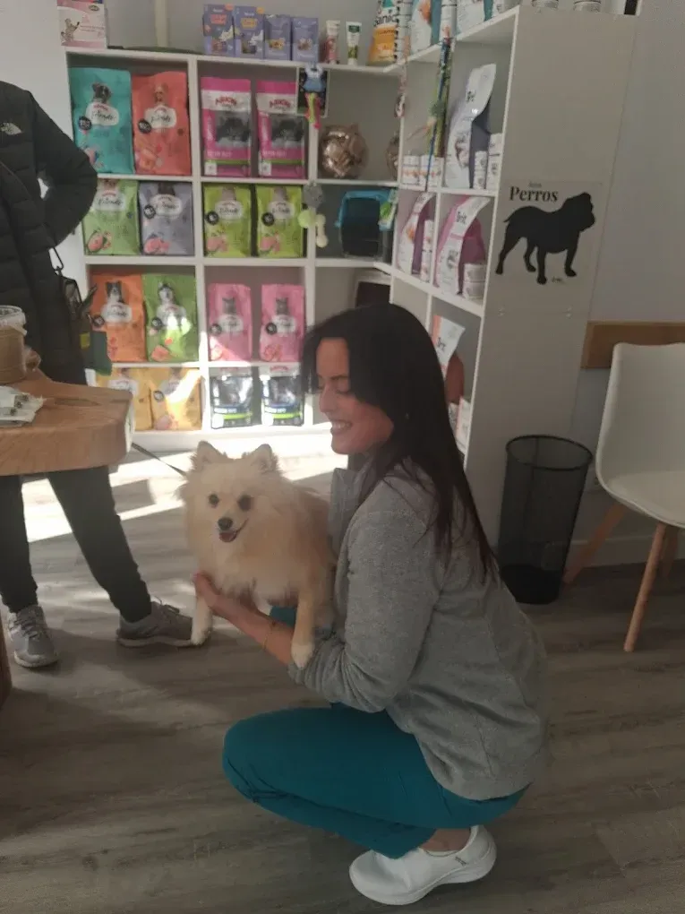 Una mujer agachada con un pequeño perro blanco en el pasillo de una tienda de mascotas, con coloridos productos para mascotas exhibidos detrás.