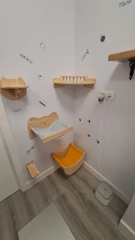 Rincón de una moderna zona de juegos para gatos con estantes montados en la pared, una hamaca y una caja de arena sobre suelo de madera.