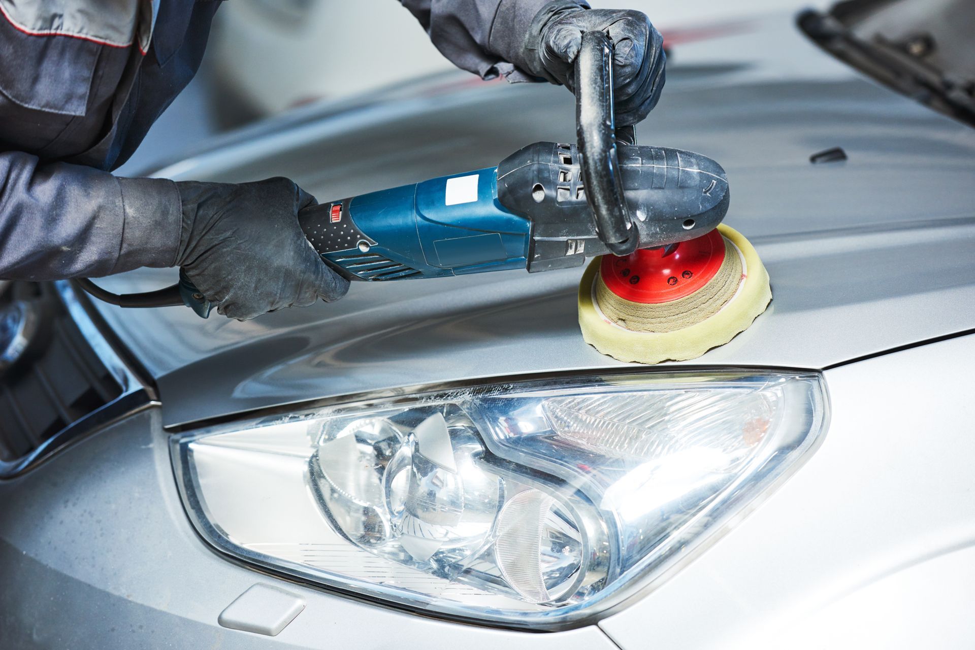 Personne polissant un phare de voiture argenté avec une polisseuse rotative, portant des gants noirs.