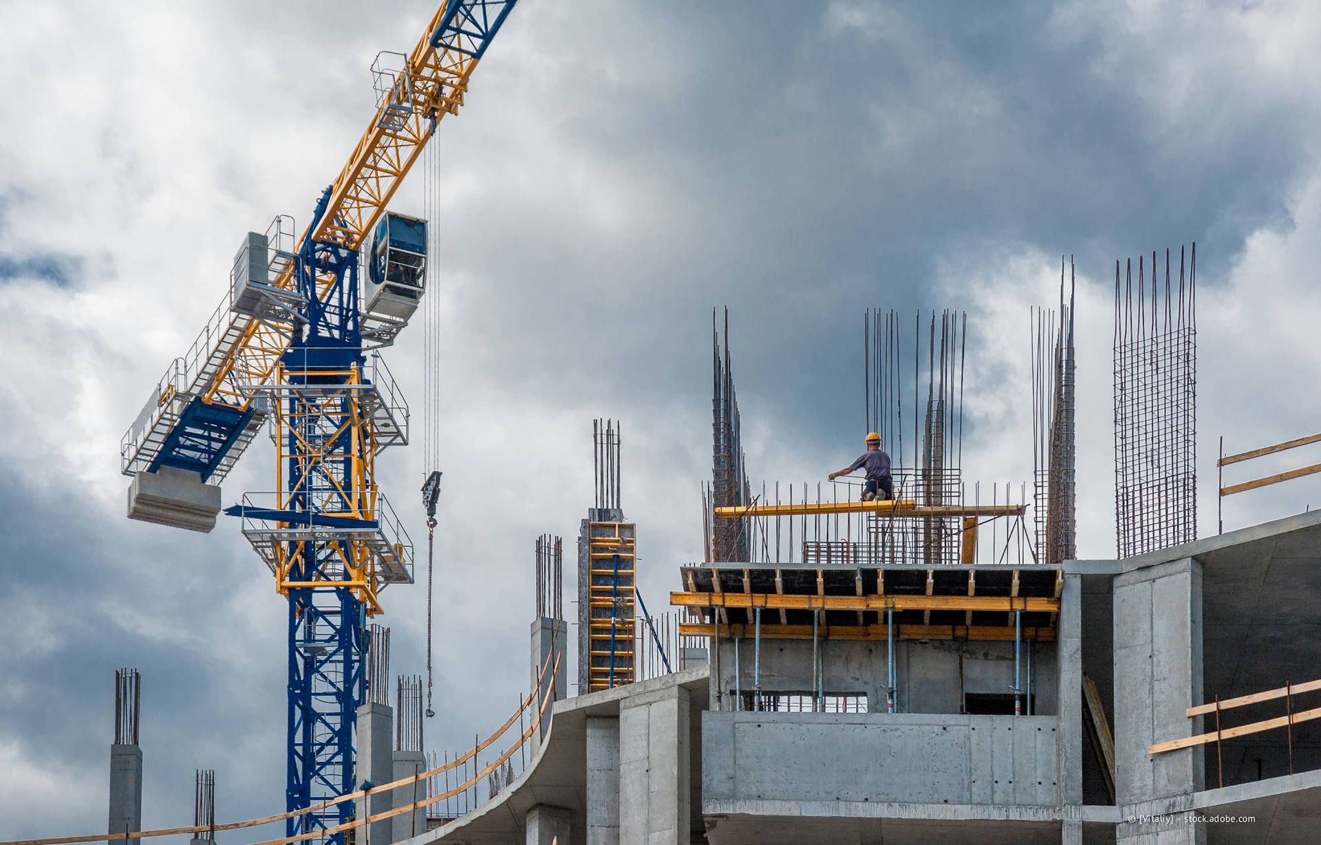 Baustelle mit Kran, der Baumaterialien hebt; Arbeiter auf Betongerüst unter bewölktem Himmel.
