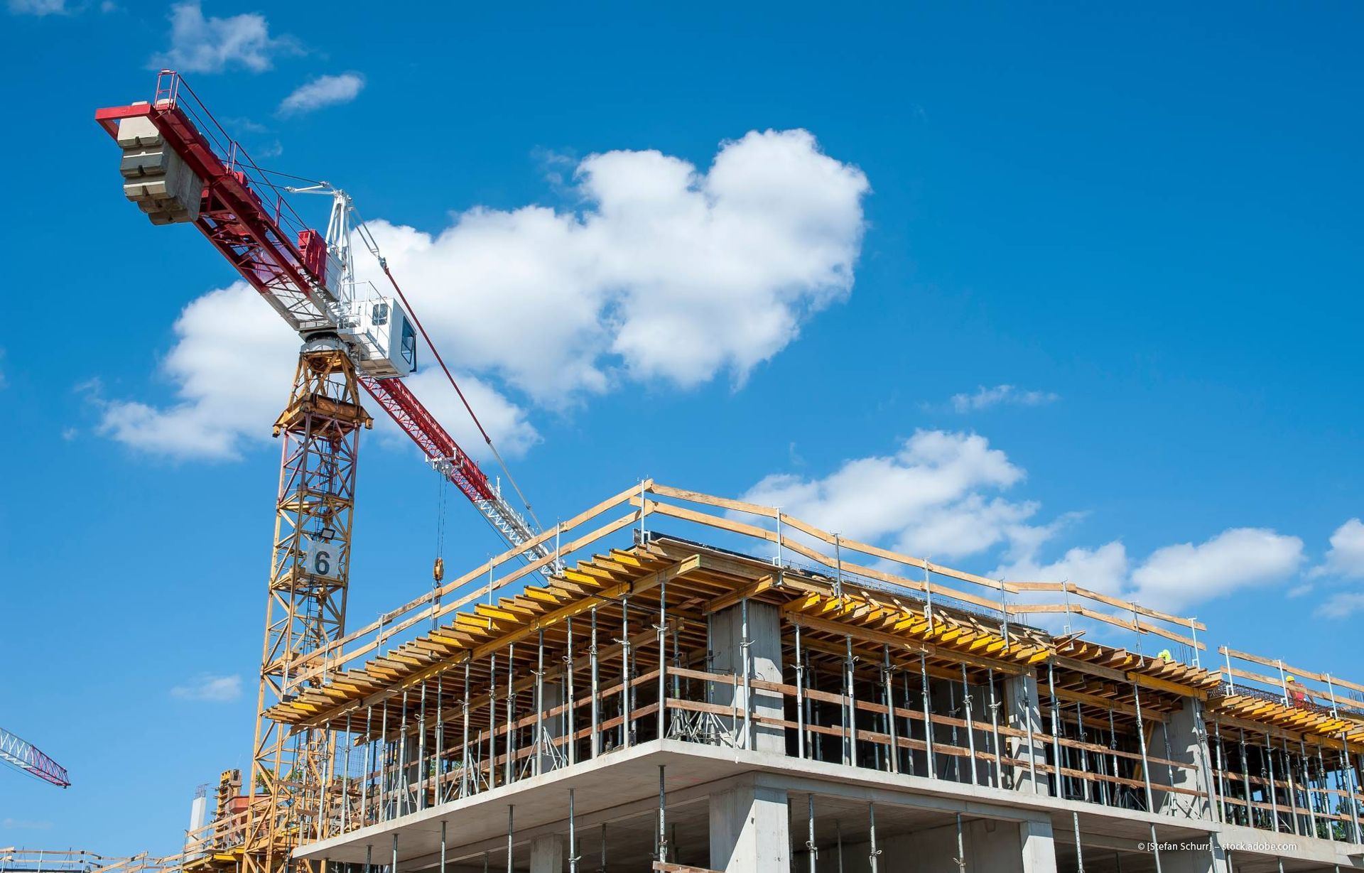 Baustelle mit Kran, der sich über ein im Bau befindliches Gebäude erstreckt; blauer Himmel und Wolken im Hintergrund.