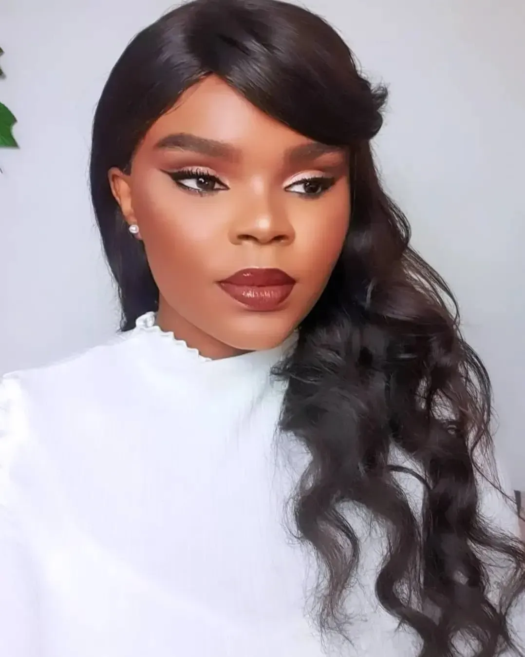 Mujer con cabello oscuro ondulado y maquillaje completo, vistiendo un top blanco, mirando ligeramente hacia un lado.