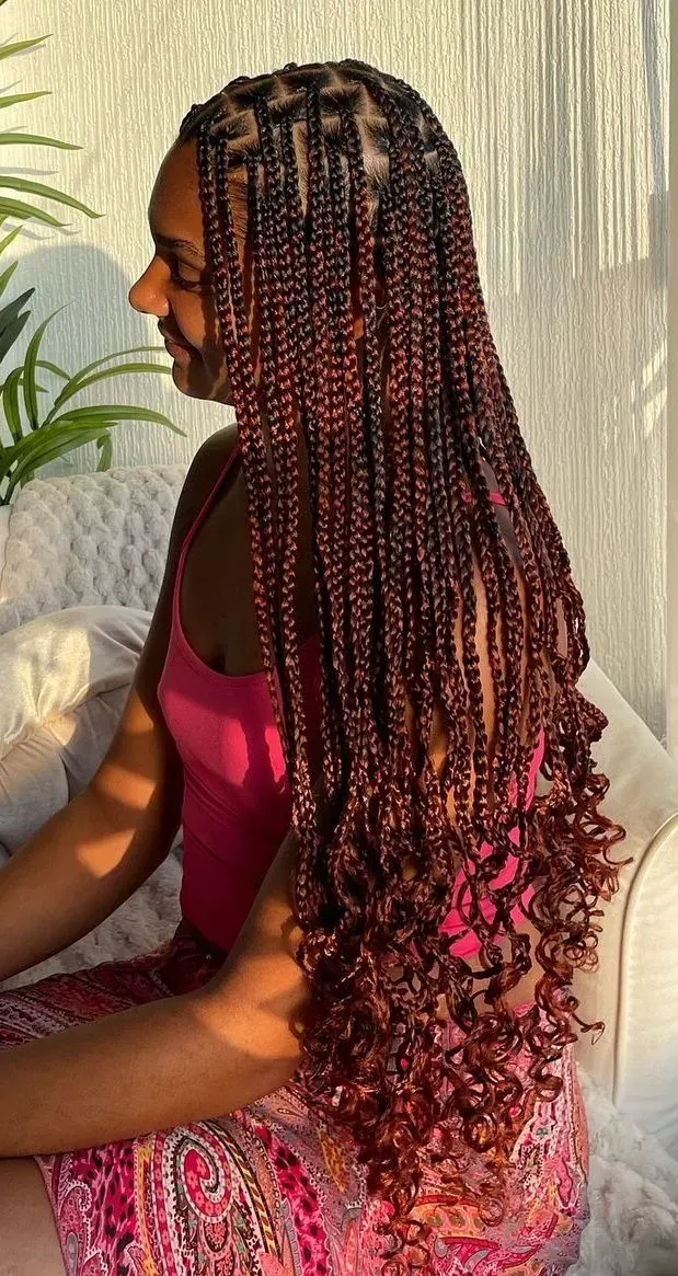 Mujer con largas trenzas de color marrón rojizo, vestida con un top rosa, sentada al aire libre bajo la luz del sol.