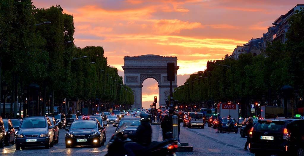 L'Arc de Triomphe encadré par les Champs-Élysées, avec une circulation dense au coucher du soleil sous un ciel aux teintes orange et rose éclatantes.