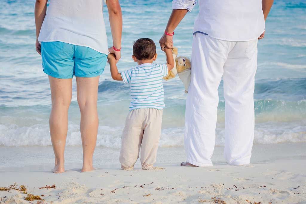 Un enfant tient la main de deux adultes, debout sur une plage de sable, face à l'océan.