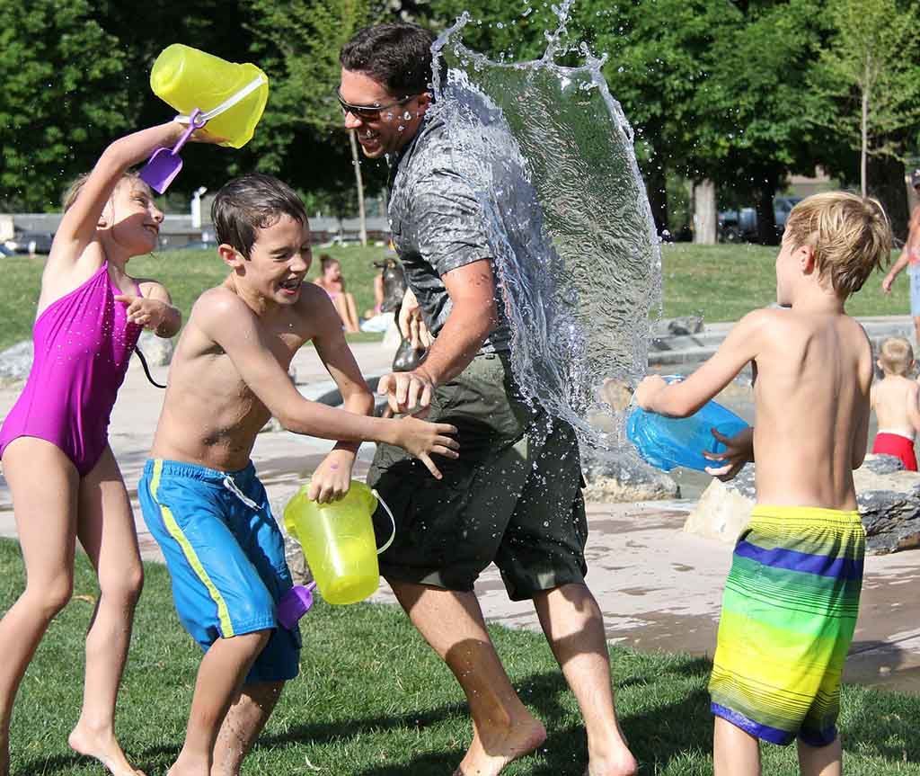 Dans un parc ensoleillé, trois enfants arrosent un homme avec des seaux d'eau.
