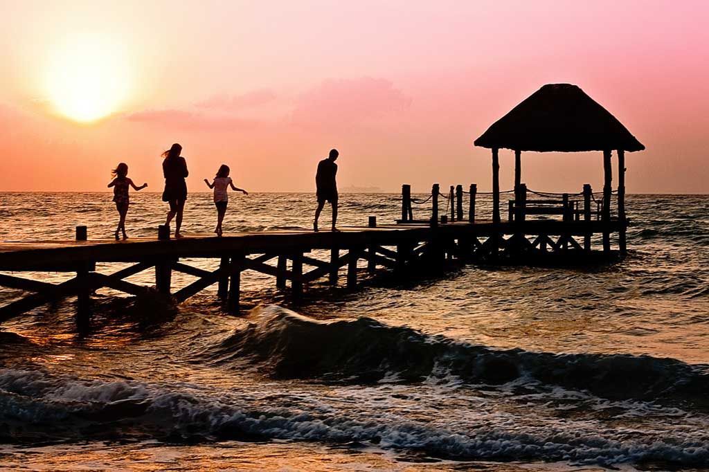 Une famille se promène sur une jetée en bois en direction d'un kiosque au coucher du soleil, sous un ciel rose et orange au-dessus de l'océan.