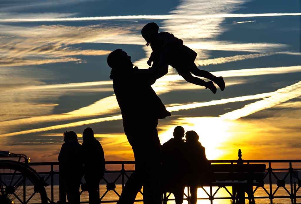 Silhouette d'une personne soulevant un enfant sur fond de coucher de soleil, près d'une balustrade, avec d'autres personnes en arrière-plan.