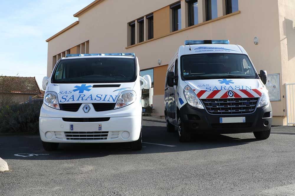 Deux ambulances blanches avec des inscriptions bleues sont stationnées devant un bâtiment.