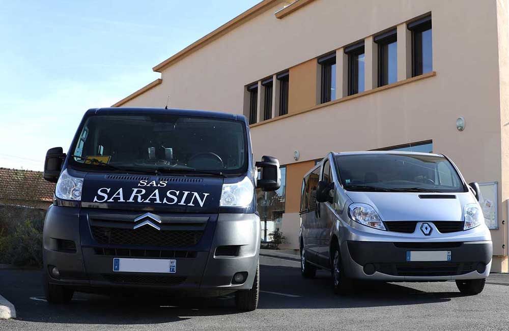 Une camionnette Citroën bleu foncé avec l'inscription SARASIN et une camionnette Renault argentée sont garées devant un bâtiment beige.
