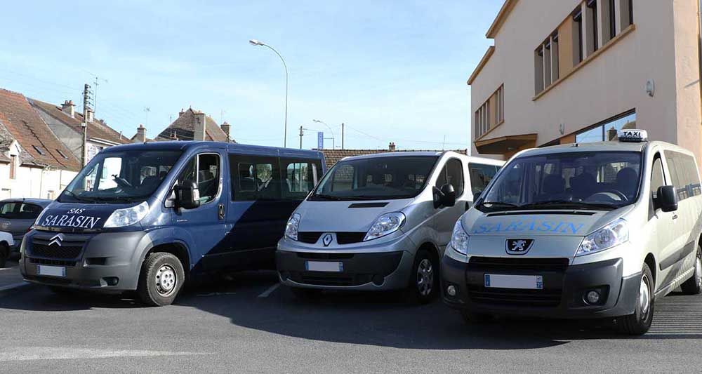 Trois fourgonnettes, de couleur bleue, argentée et grise, étaient garées côte à côte devant un bâtiment par une journée ensoleillée.