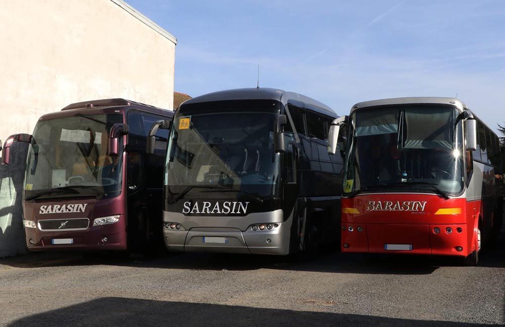 Trois bus touristiques Sarasin — un bordeaux, un gris et un rouge — étaient garés en rang à l'extérieur par une journée ensoleillée.