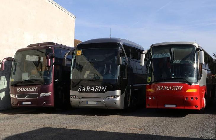 Trois bus touristiques Sarasin — un bordeaux, un gris et un rouge — étaient garés en rang à l'extérieur par une journée ensoleillée.