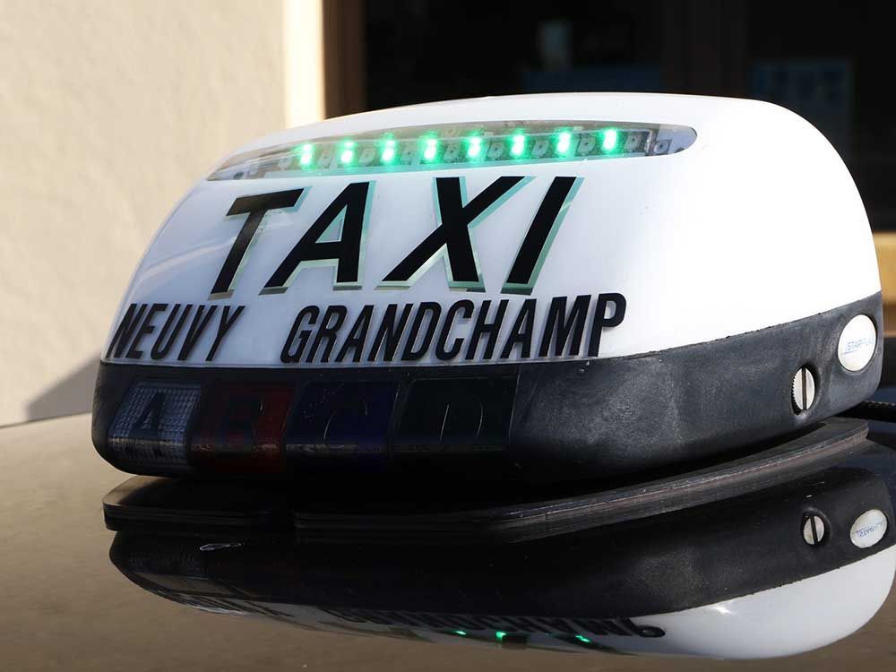 Un panneau blanc sur le toit d'un taxi, avec des lumières vertes indiquant TAXI et NEUVY GRANDCHAMP, sur une surface sombre et réfléchissante.
