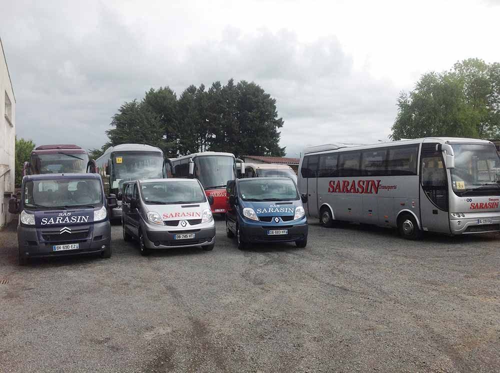 Une flotte de fourgonnettes et d'autobus gris et blancs de la marque Sarasin est stationnée sur un terrain gravillonné sous un ciel couvert.