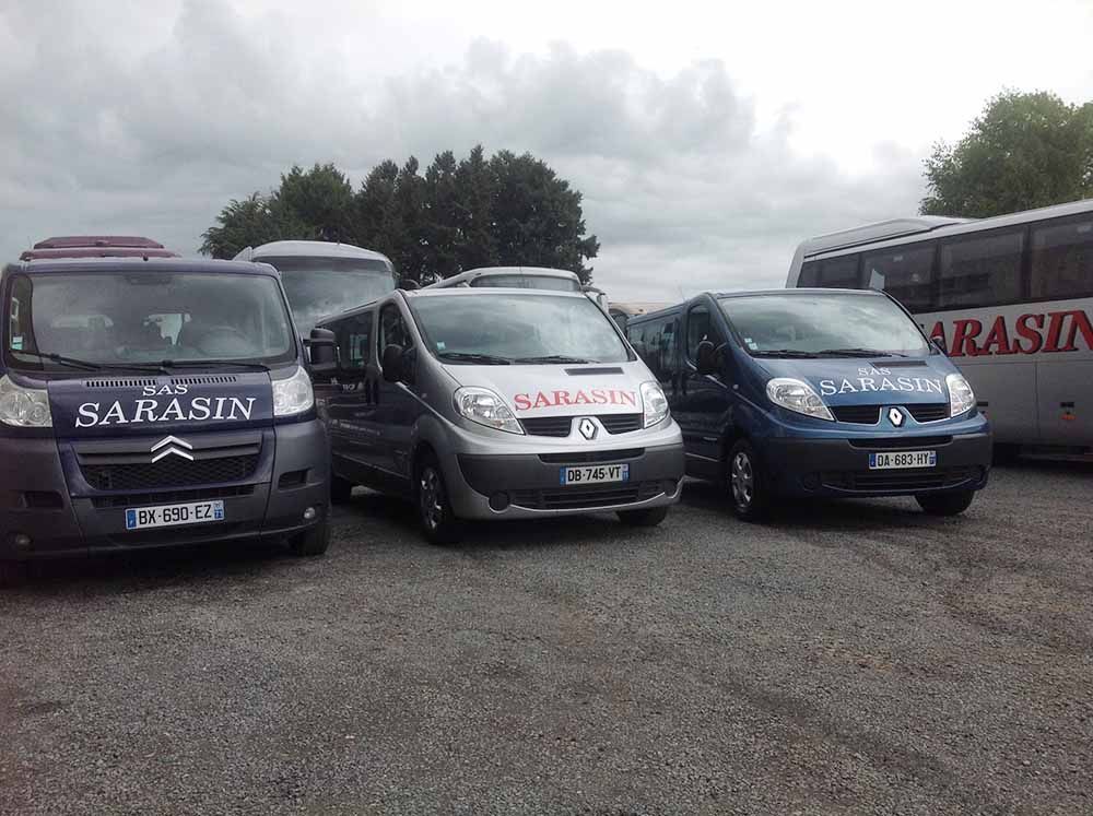 Une flotte de véhicules de marque Sarasin, comprenant trois fourgonnettes et un autobus, était stationnée sur un terrain gravillonné sous un ciel nuageux.
