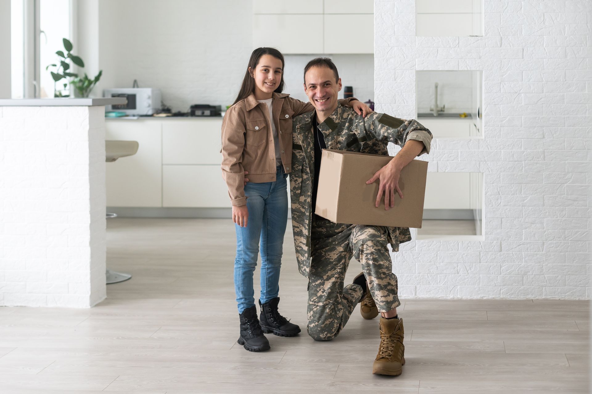 Un militaire et une enfant souriants posent dans une cuisine lumineuse, l'homme tenant une boîte en carton.