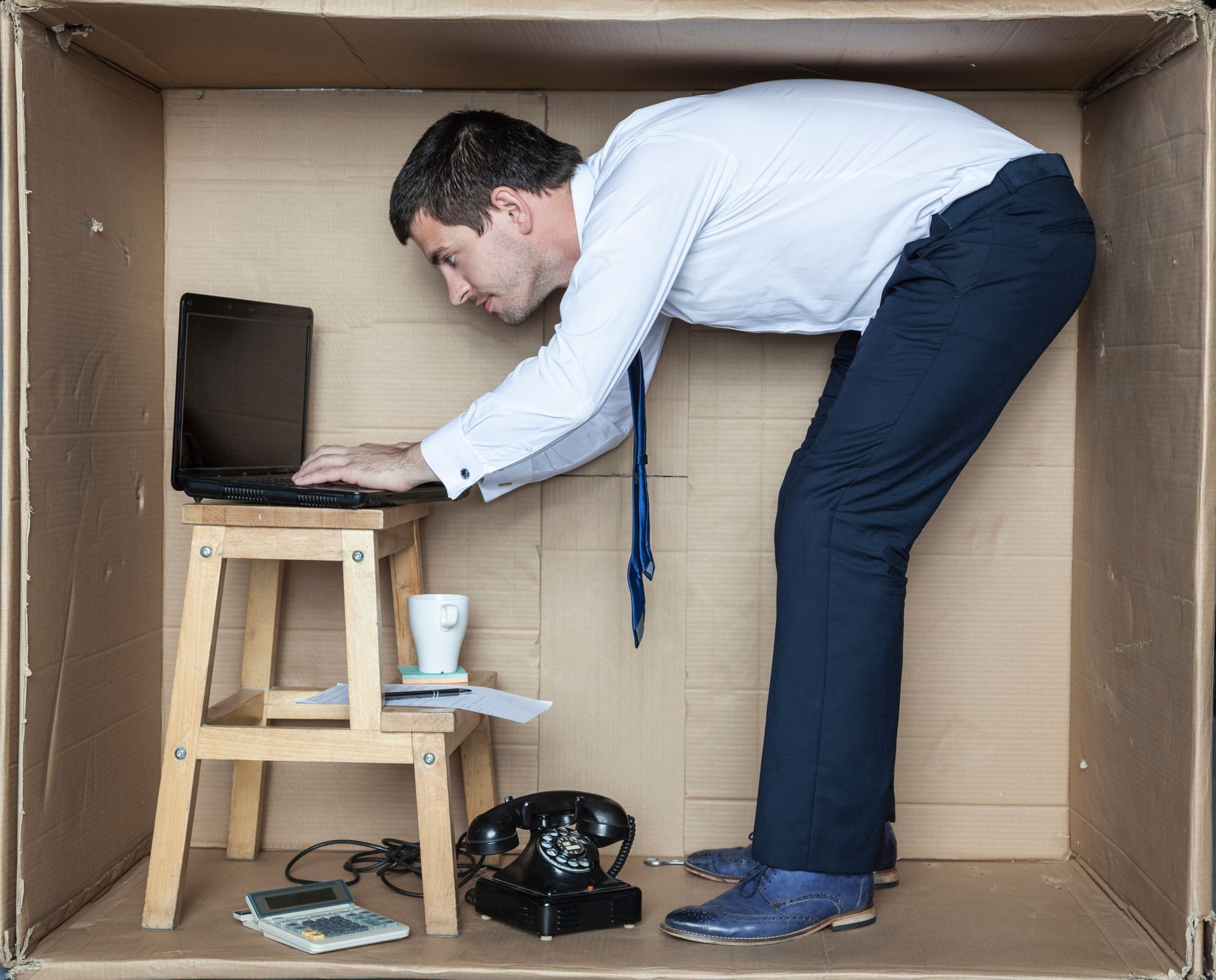 Un homme penché dans sur un énorme carton, tapant sur un ordinateur portable à côté d'un tabouret et d'un téléphone