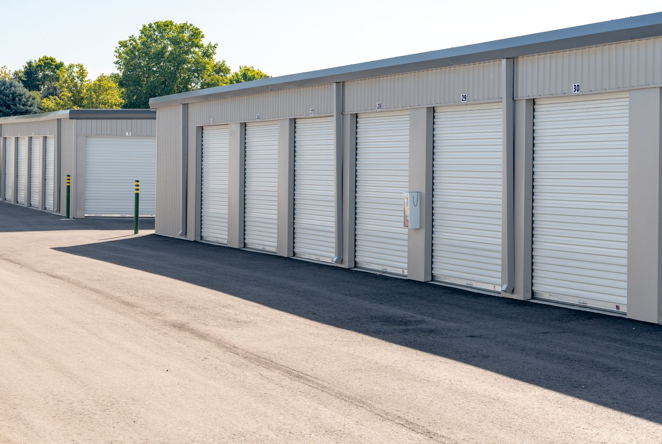 Une rangée de portes de box de stockage longe un terrain pavé, à côté d'un bâtiment beige.