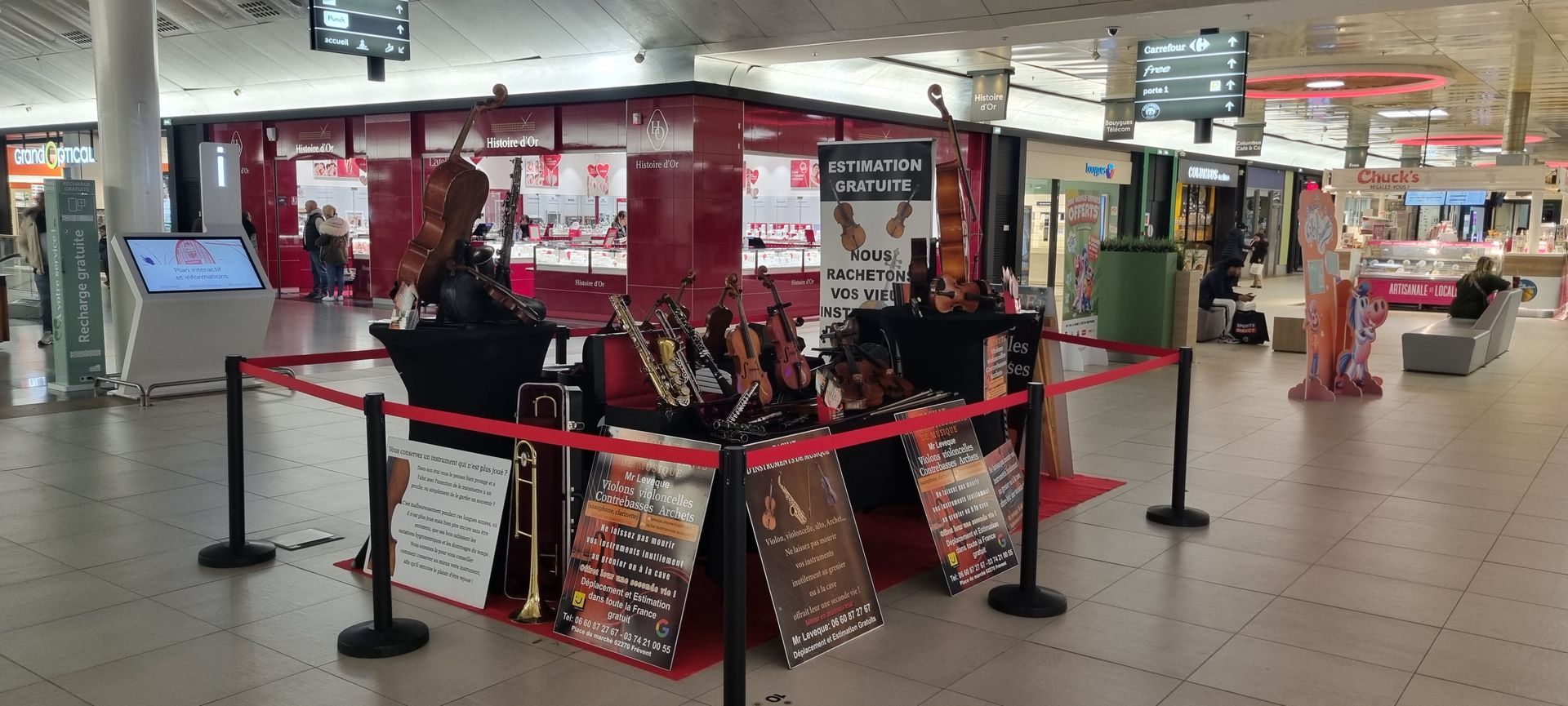 Une exposition d'instruments de musique dans un centre commercial, présentant notamment des violons, est entourée de barrières et de panneaux rouges.