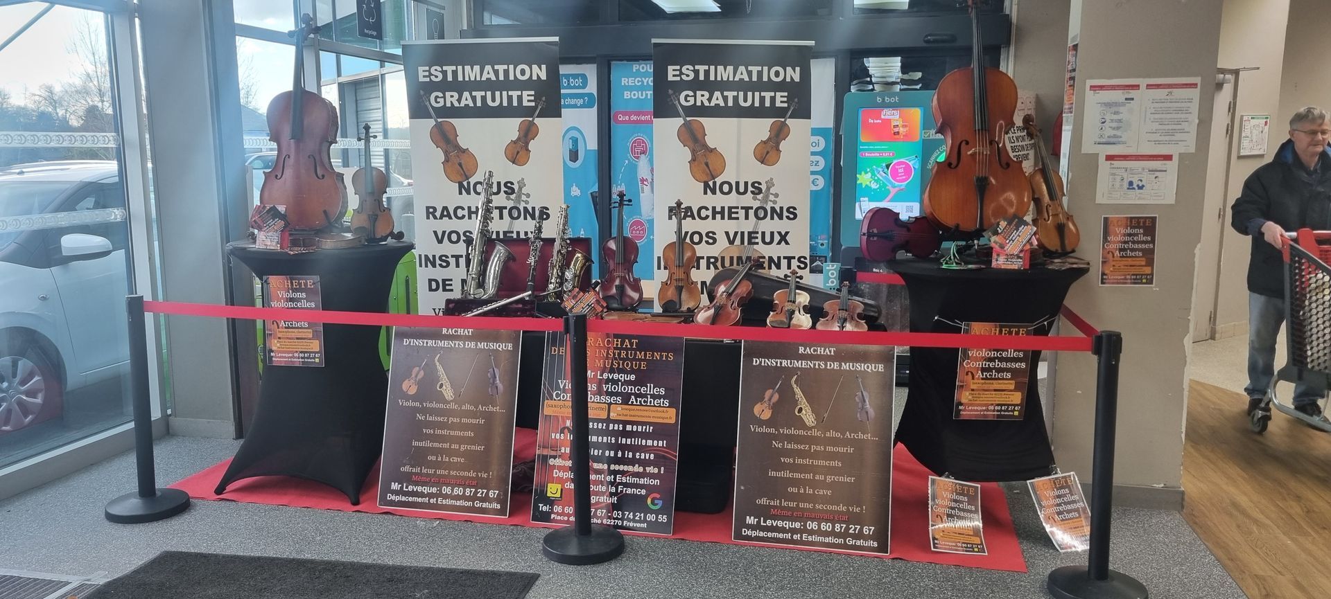 Vitrine de magasin avec violons et panneaux publicitaires. Barrière rouge devant la vitrine. Une personne passe sur la droite.