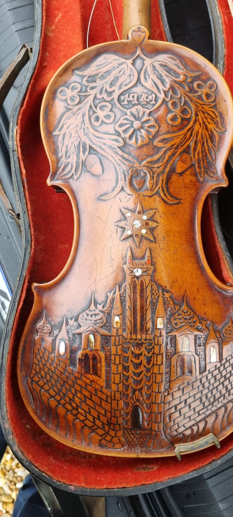 Le fond du violon est finement sculpté de motifs floraux et d'un bâtiment. Le violon est rangé dans son étui doublé de rouge.