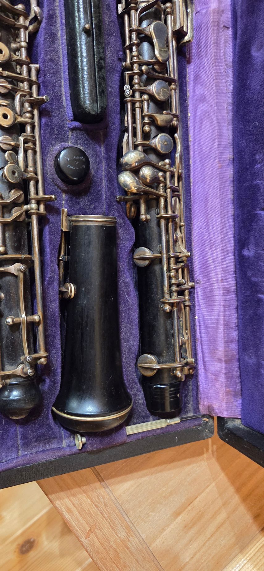 Un hautbois dans son étui, avec ses pièces et ses clés métalliques, posé sur un plancher en bois. L'intérieur de l'étui est recouvert de velours violet.