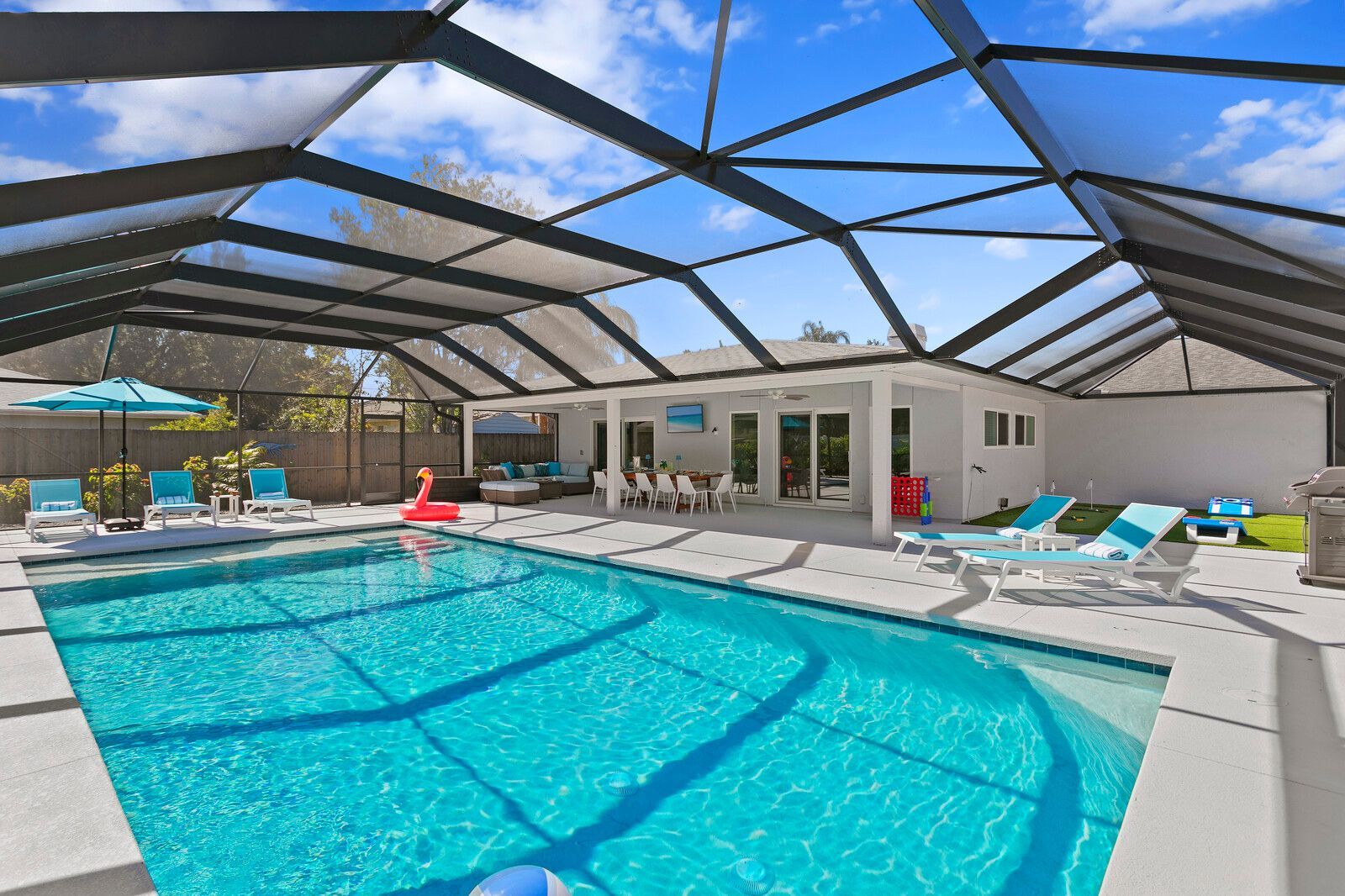 Swimming pool under a screened enclosure with lounge chairs, a dining area, and a white house on a sunny day.