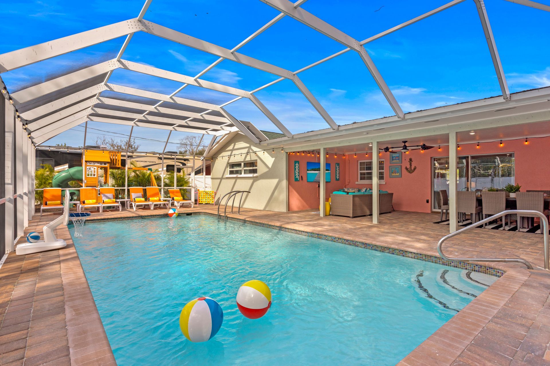 Pool with a screened enclosure, beach balls floating, chairs, and a covered seating area in a sunny backyard.