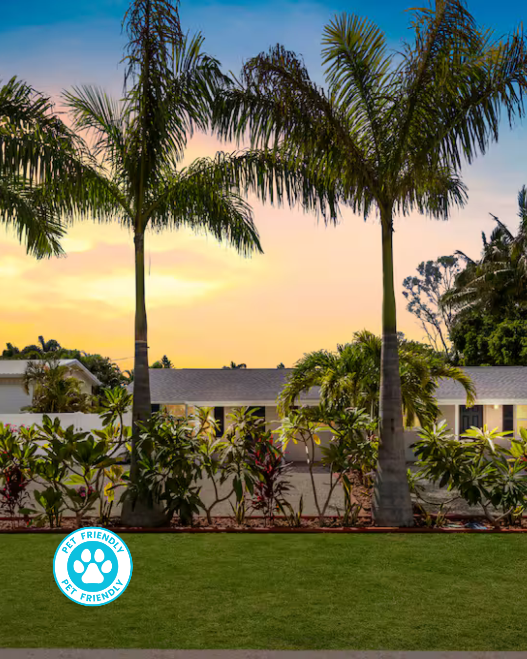 Palm trees in front of a light yellow house at sunset, with a green lawn and driveway.