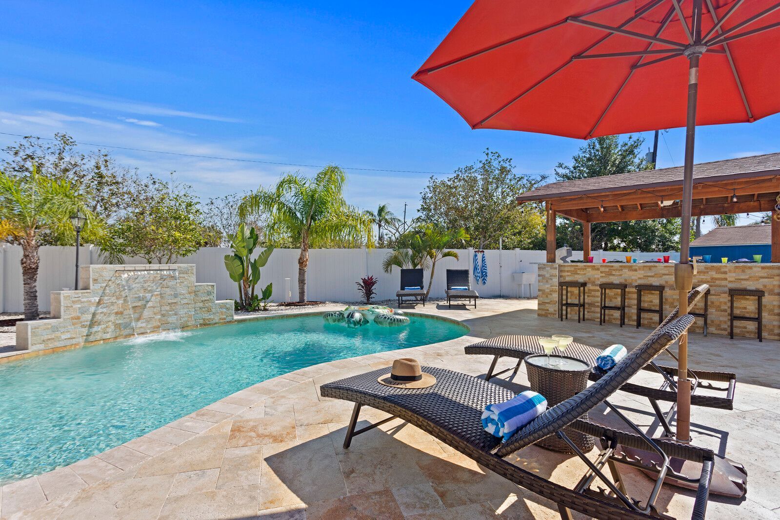 Swimming pool with waterfall, lounge chairs, and bar under a red umbrella on a sunny day.