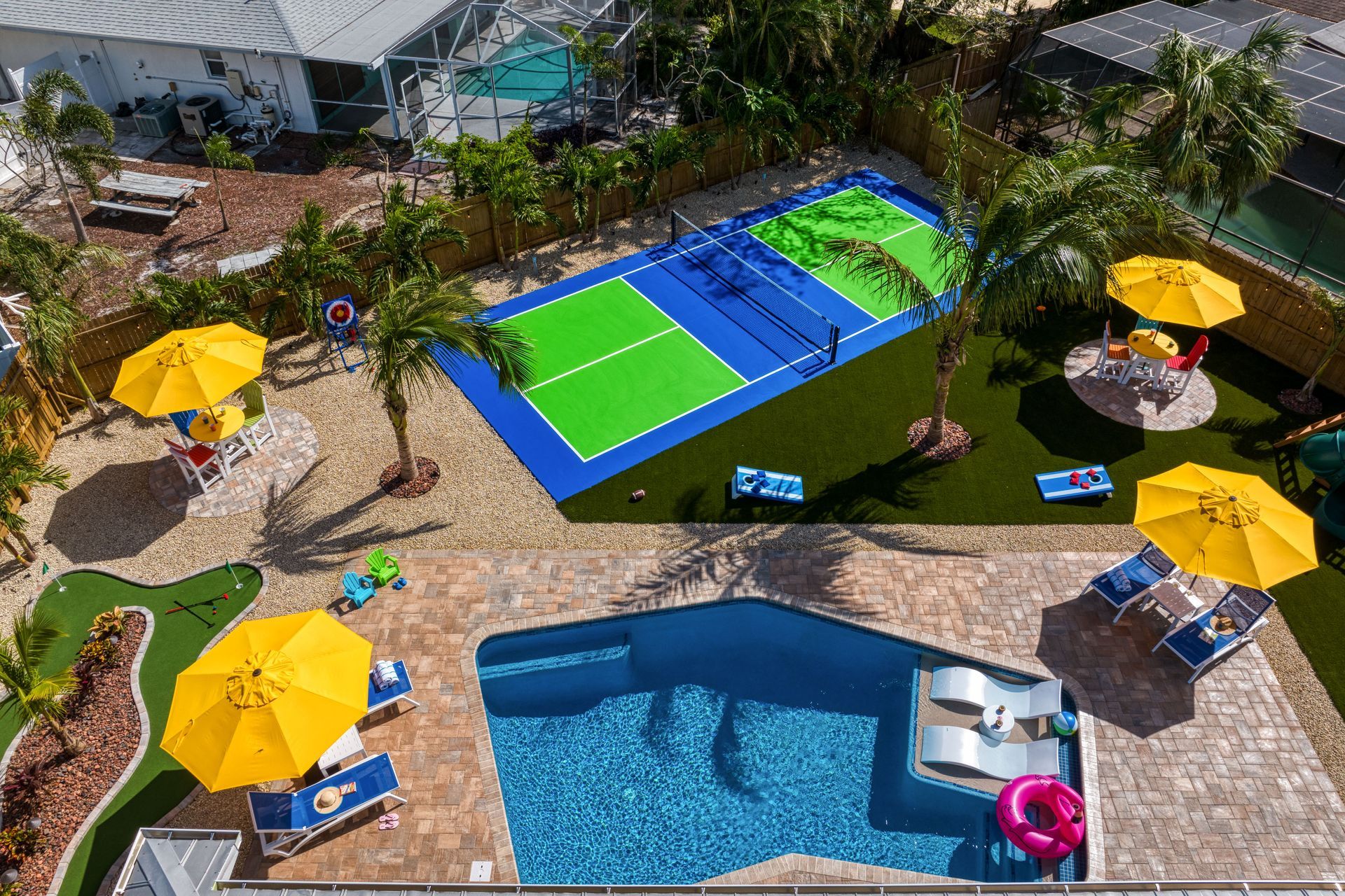Aerial view of a backyard with a pool, pickleball court, and putting green, with yellow umbrellas and palm trees.