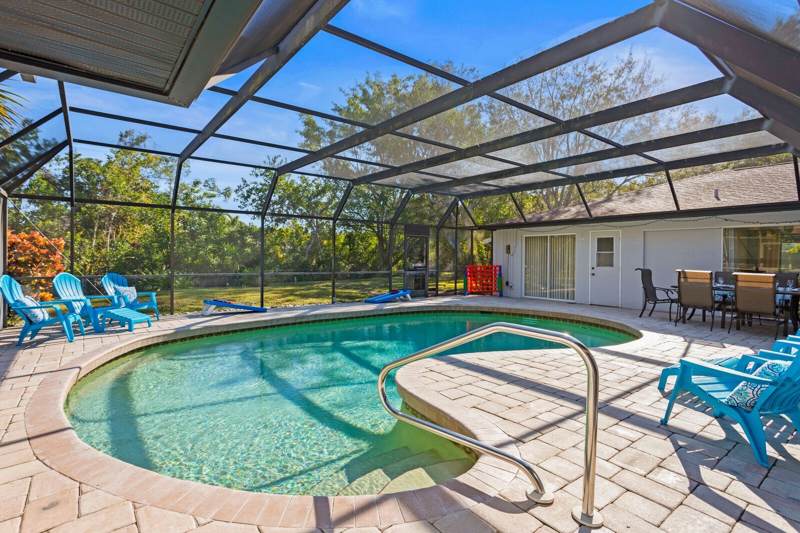 Enclosed backyard pool with blue chairs, a dining area, and a screened enclosure under a bright blue sky.