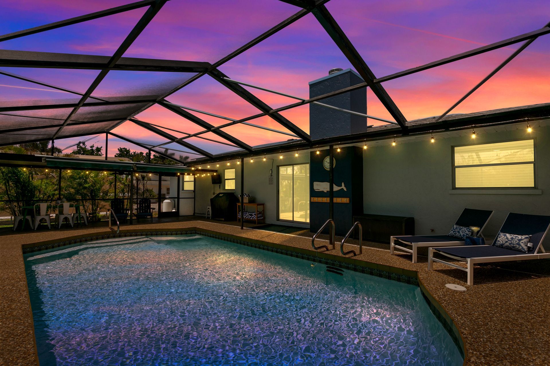 Pool area at dusk with pool, lounge chairs, string lights, and colorful sky.