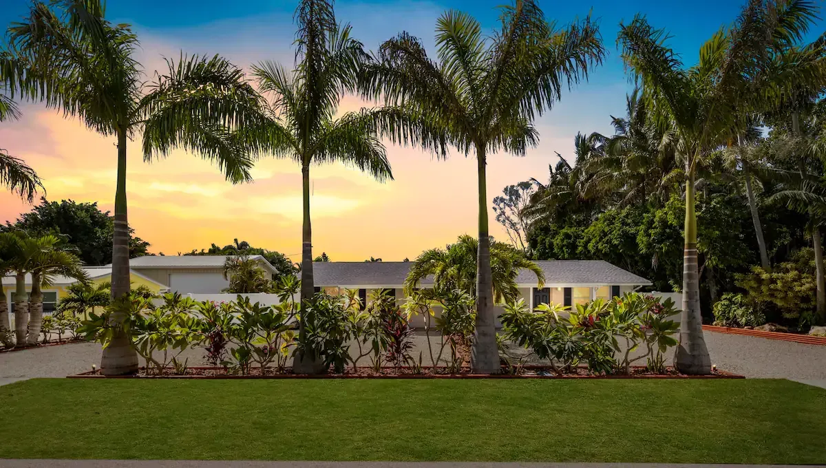 Palm trees in front of a light yellow house at sunset, with a green lawn and driveway.