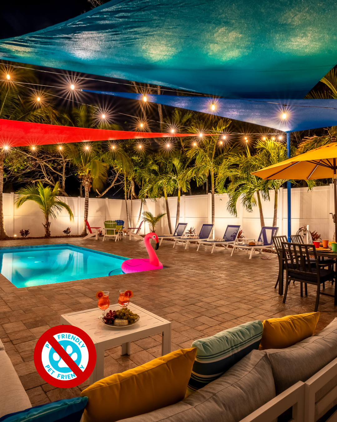 Poolside patio at night, featuring pool, string lights, colorful shade sails, seating, and a grill.
