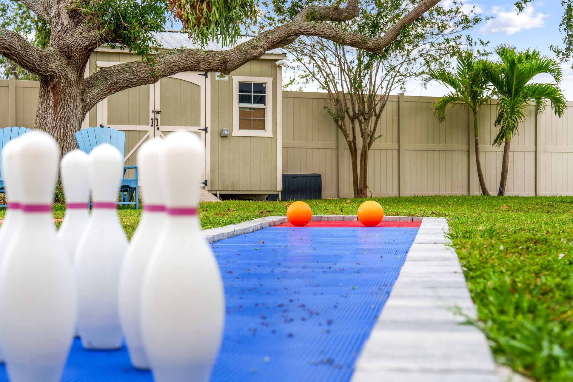 Bowling setup outdoors: white pins, orange balls, blue lane, grassy yard.