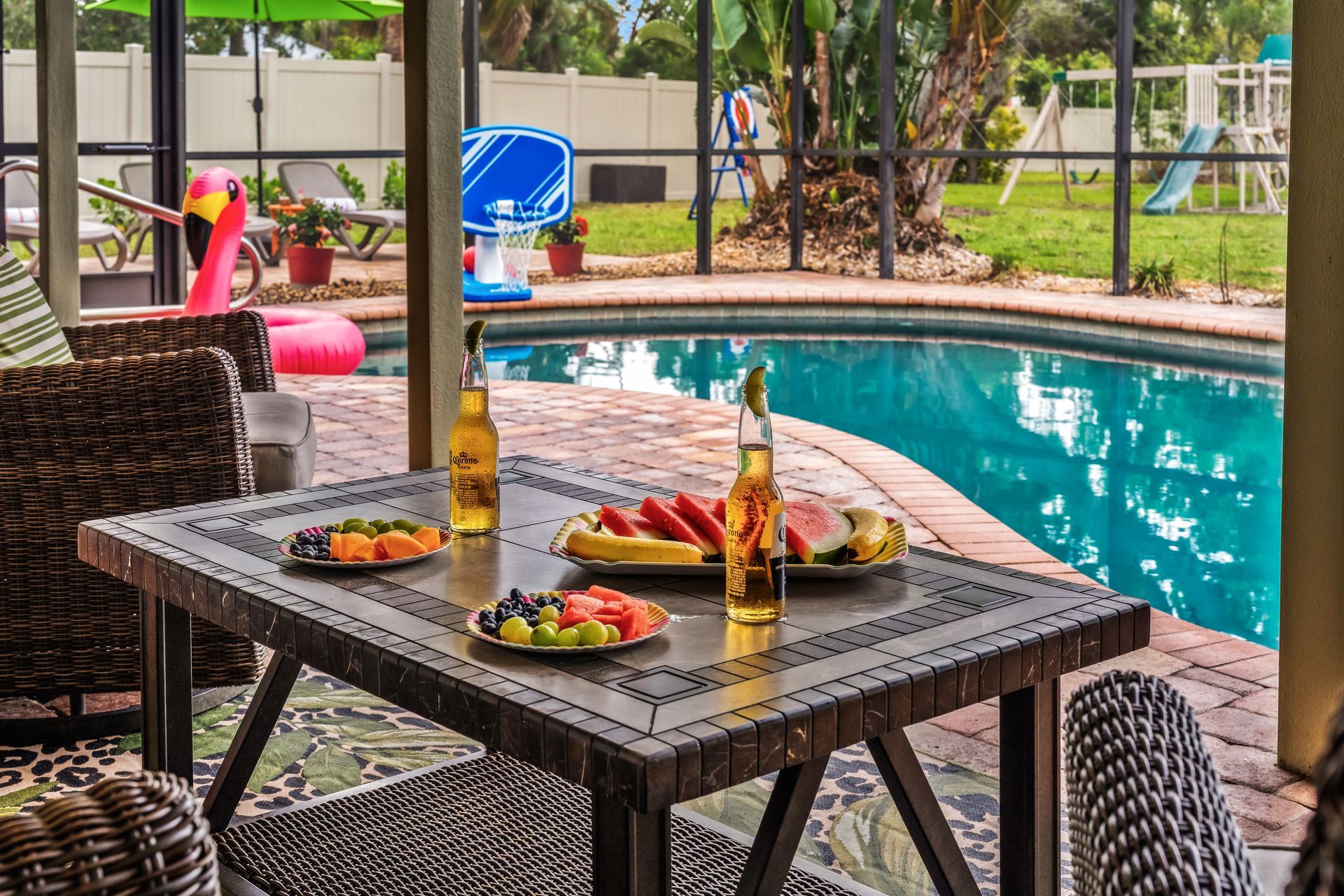A table with fruit and beer near a pool. Backyard with a flamingo floatie and a playground.
