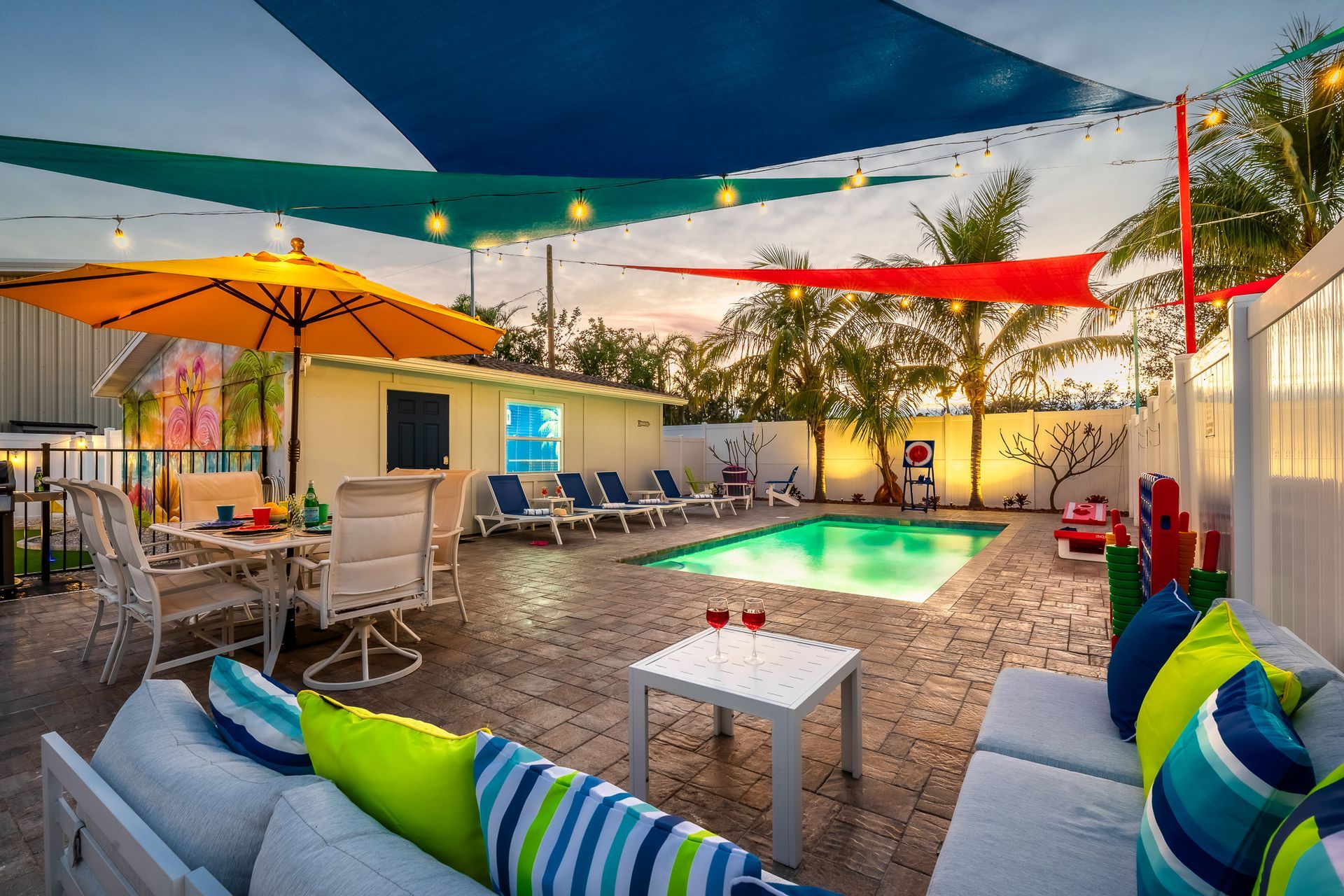 Poolside patio with white furniture, colorful pillows, and shade sails.