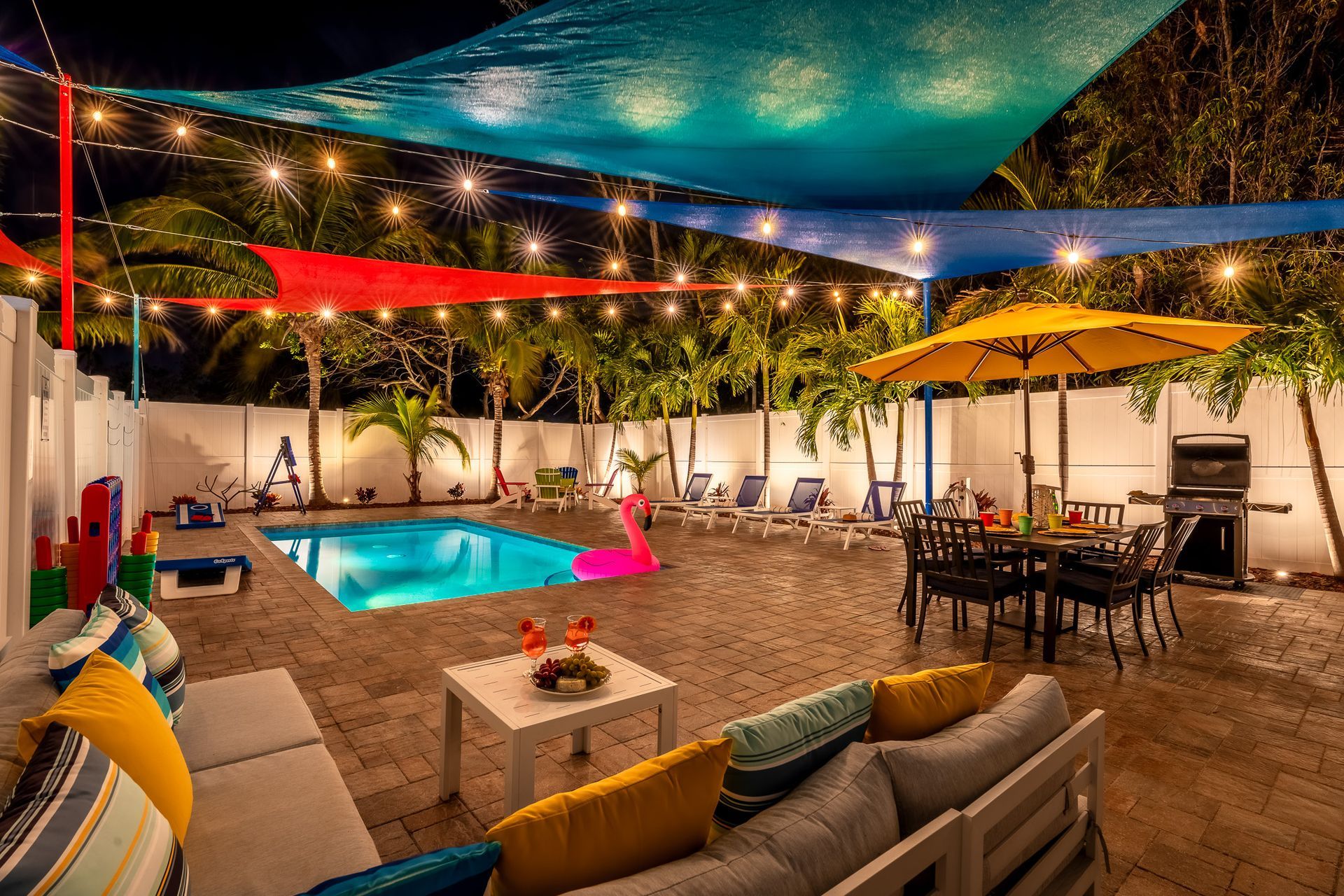 Poolside patio at night, featuring pool, string lights, colorful shade sails, seating, and a grill.