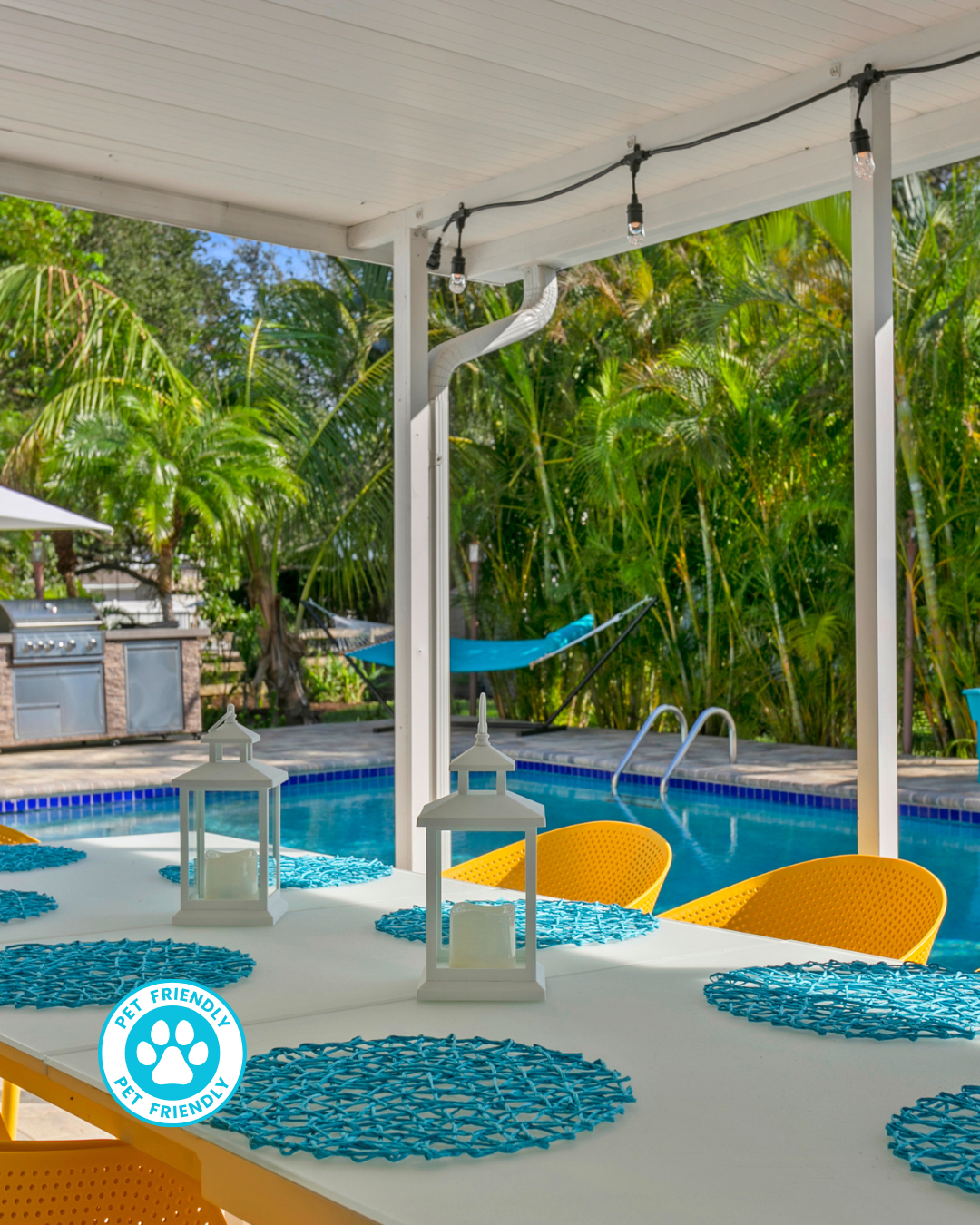 Poolside patio with a dining table, chairs, and outdoor grill. Blue pool and lounge chairs, lush greenery in the background.