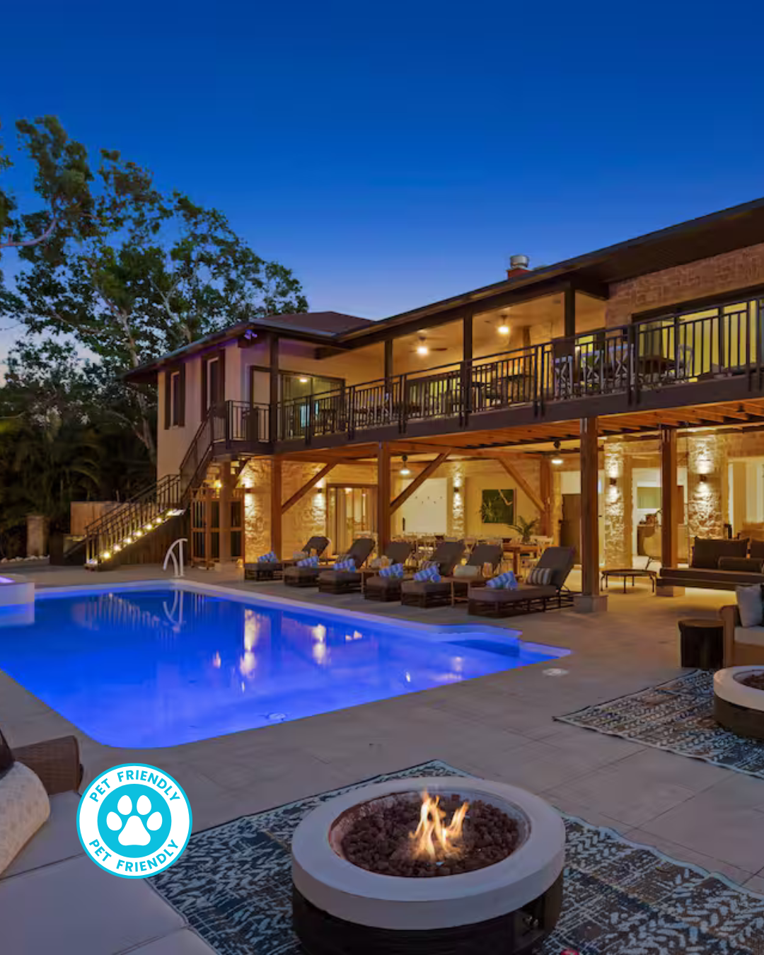 A two-story house at twilight with a lit swimming pool, lounge chairs on the patio, and a glowing outdoor fire pit.