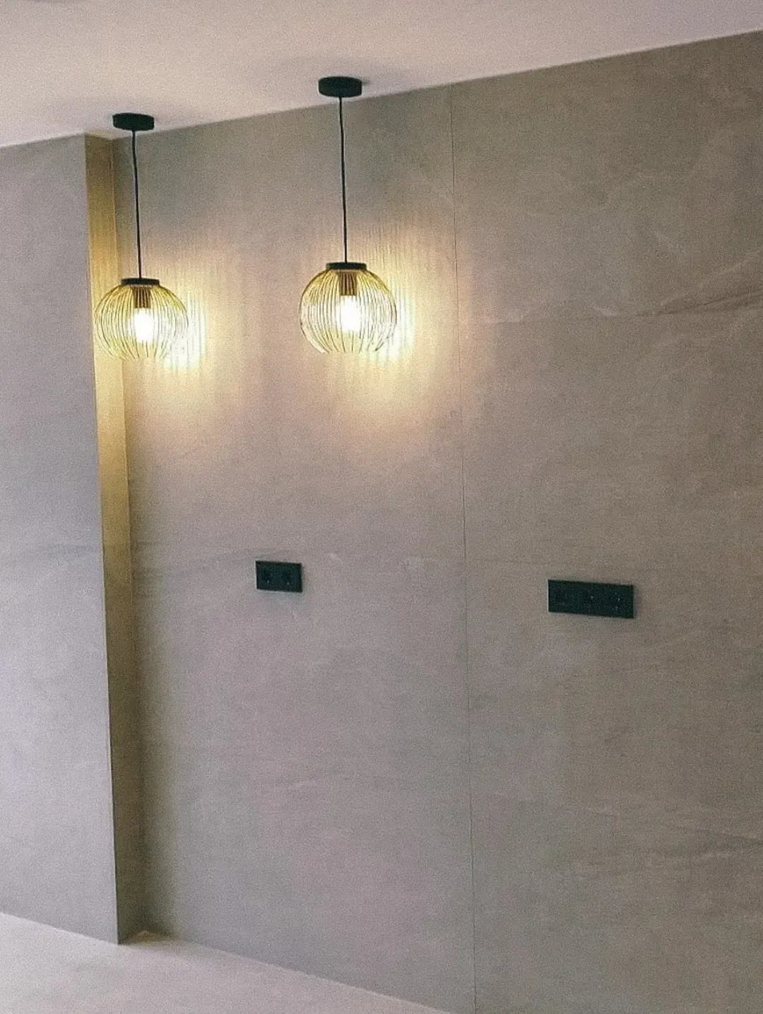 Dos lámparas colgantes iluminan una pared de azulejos grises, con dos tomas de corriente oscuras debajo.