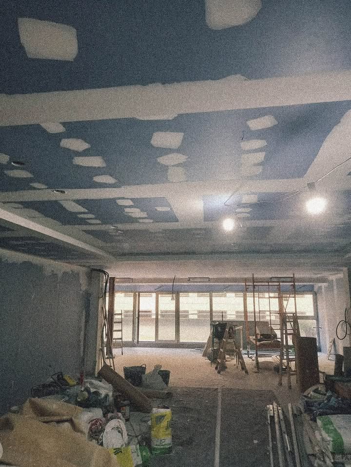Interior de obra con techo azul, paneles de yeso parchados y paredes sin terminar. Vista hacia la fachada acristalada.